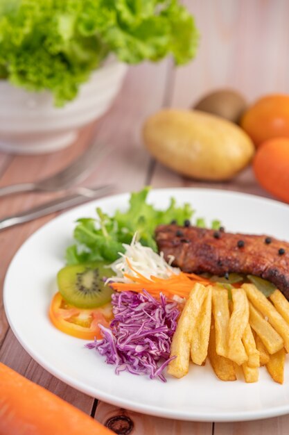 Vis steak met frietjes, kiwi, sla, wortelen, tomaten en kool in een witte schotel.