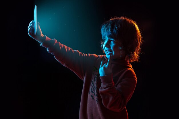 Vloggen met smartphone. Portret van een blanke jongen op een donkere muur in neonlicht. Prachtig model met krullen. Concept van menselijke emoties, gezichtsuitdrukking, verkoop, advertentie, moderne technologie, gadgets.