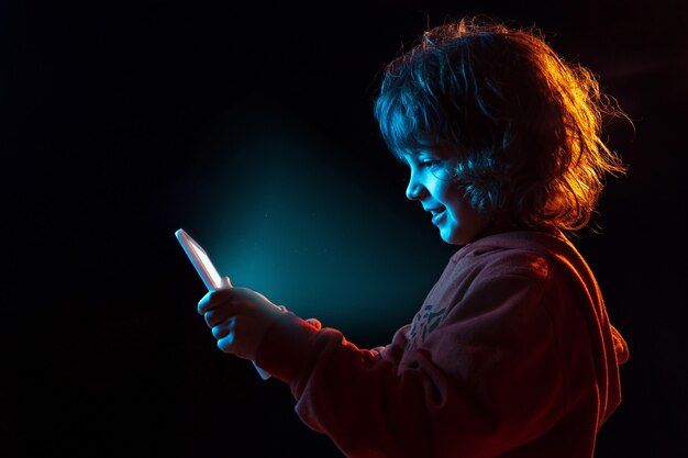 Vloggen met tablet, spelen. Het portret van de Kaukasische jongen op donkere studioachtergrond in neonlicht. Prachtig krullend model. Concept van menselijke emoties, gezichtsuitdrukking, verkoop, advertentie, moderne technologie, gadgets.