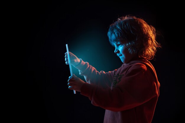 Vloggen met tablet, spelen. Het portret van de Kaukasische jongen op donkere studioachtergrond in neonlicht. Prachtig krullend model. Concept van menselijke emoties, gezichtsuitdrukking, verkoop, advertentie, moderne technologie, gadgets.