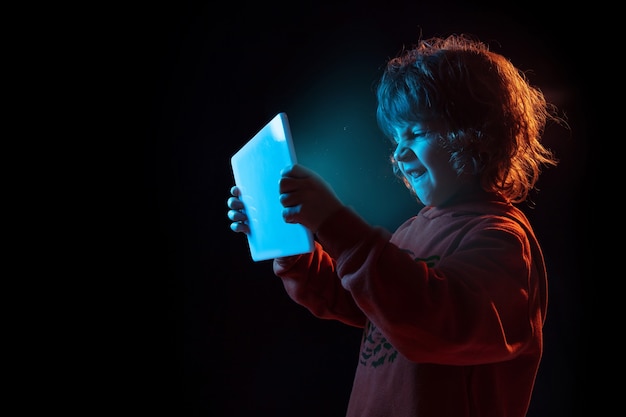 Vloggen met tablet, spelen. Portret van een blanke jongen op een donkere muur in neonlicht. Prachtig krullend model. Concept van menselijke emoties, gezichtsuitdrukking, verkoop, advertentie, moderne technologie, gadgets.