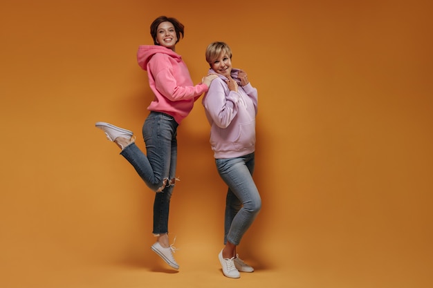 Volle lengte foto van jonge dame in roze hoodie en skinny jeans springen en lachend samen met oude vrouw op oranje achtergrond.