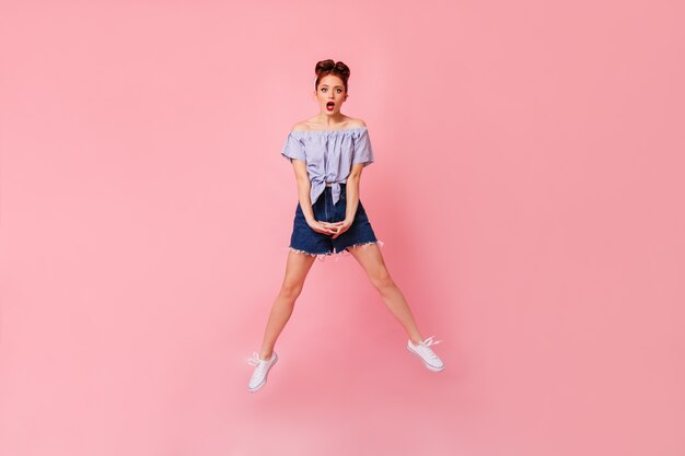 Volle lengte weergave van geschokte vrouw in denim shorts en blouse. Studio shot van verbaasd pinup meisje springen op roze ruimte.