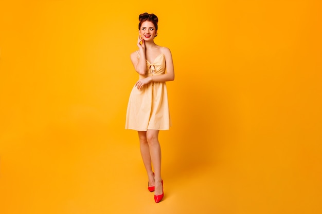 Volle lengte weergave van vrolijke pinup dame. Aantrekkelijke jonge vrouw in jurk staande op gele ruimte.
