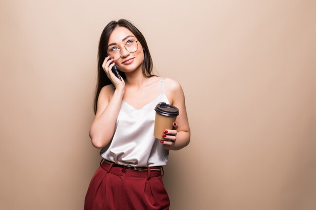 Volledig lengteportret van een glimlachende Aziatische mobiele telefoon van de vrouwenbespreking terwijl het houden van kop van koffie geïsoleerd over beige muur gaan