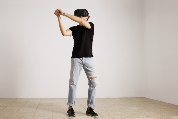 Volledig lengteportret van een jong Kaukasisch model in lichtblauwe gescheurde jeans en zwart t-shirt die honkbal of tennis spelen in VR-bril