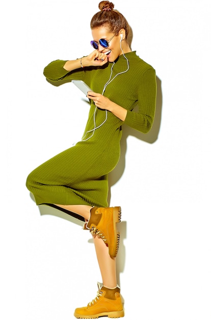 Volledig lengteportret van mooi gelukkig leuk glimlachend donkerbruin vrouwenmeisje in de toevallige groene kleren van de hipsterzomer die op witte het luisteren muziek in smartphone met hoofdtelefoons wordt geïsoleerd