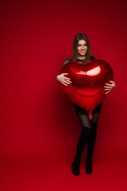 Volledig lengteportret van vrolijk donkerbruin meisje in kleding en laarzen die rode hartvormige ballon op rode achtergrond omhelzen. Sint Valentijn concept.