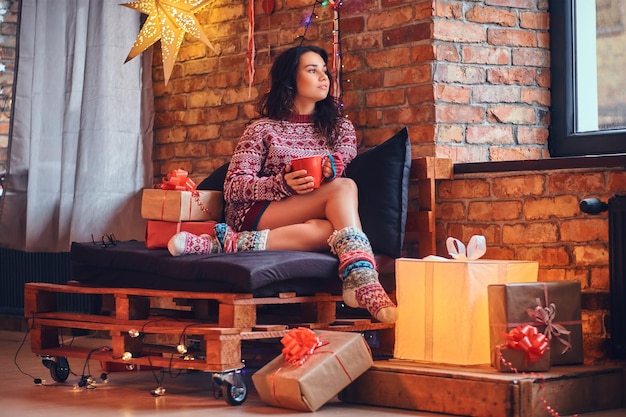 Volledig lichaamsbeeld van een sexy brunette vrouw drinkt koffie in een kamer met kerstversiering.