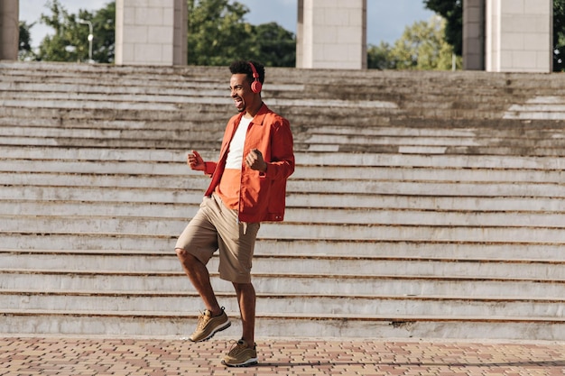 Volledig portret van brunet man in beige korte broek oranje jas en kleurrijke tshirt dansen en luisteren naar muziek in rode koptelefoon buiten