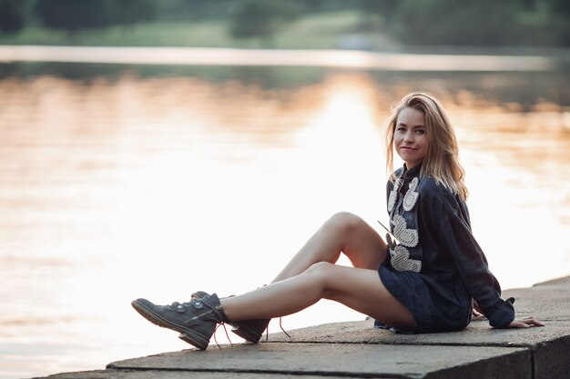 Volledig portret van een mooi blond Kaukasisch meisje in donkergrijze casual hoodiejurk en laarzen zittend bij de rivier bij zonsondergang die zonlicht op het oppervlak werpt...