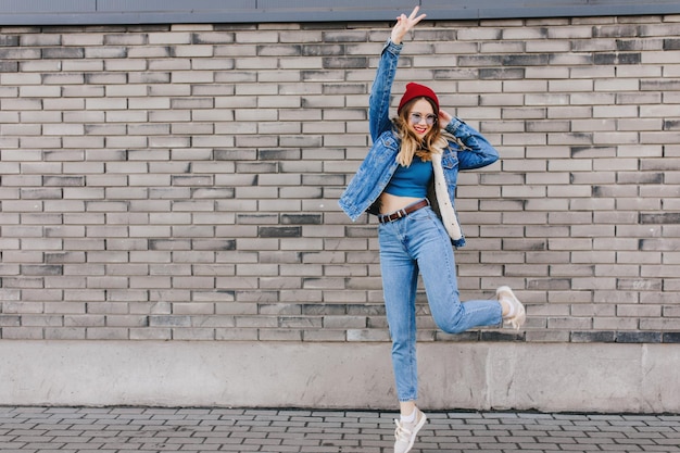 Volledige foto van betoverende dame in sportschoenen en spijkerjasje dansen in de buurt van bakstenen muur buiten schot van kaukasisch meisje springen op straat met blije gezichtsuitdrukking