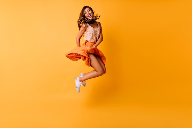 Volledige foto van gebruind gembermeisje dat op gele achtergrond springt en glimlacht Schattige jonge vrouw in trendy zomerkleren die in de studio voor de gek houden