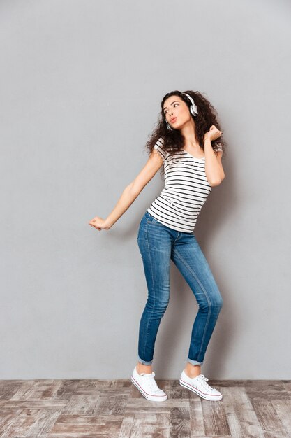 Volledige groottemening van charmante jonge vrouw in gestreepte t-shirt en jeans die terwijl het luisteren melodieën via hoofdtelefoons over grijze muur dansen