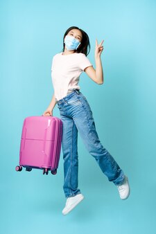 Volledige lengte van gelukkige aziatische vrouw die van vreugde springt met koffer, medisch gezichtsmasker draagt van covid, enthousiast over vakantie in het buitenland, toerisme en gezondheidszorgconcept.