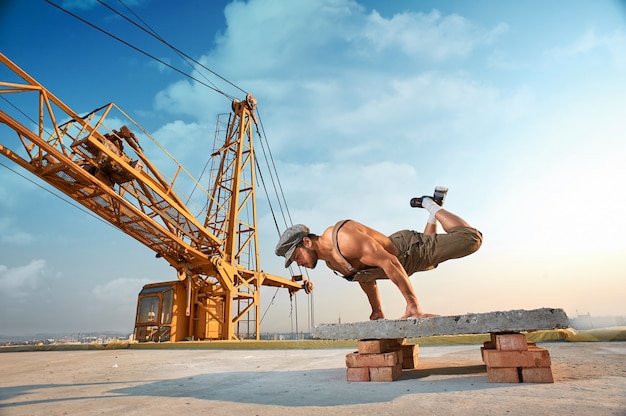 Volledige lengte van gespierde en atletische man die oefeningen doet bij handen en push-ups op handen. Un afwerking gebouw op hoog. Grote ijzeren kraan op de achtergrond.