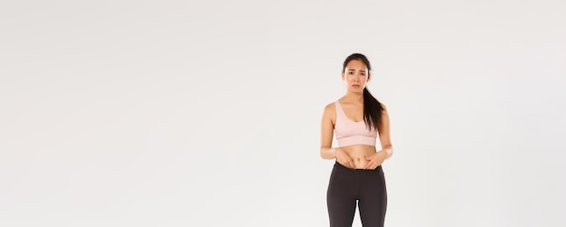 Volledige lengte van overstuur en somber aziatisch meisje in fitnesskleding die vet op de buik toont en klaagt over b