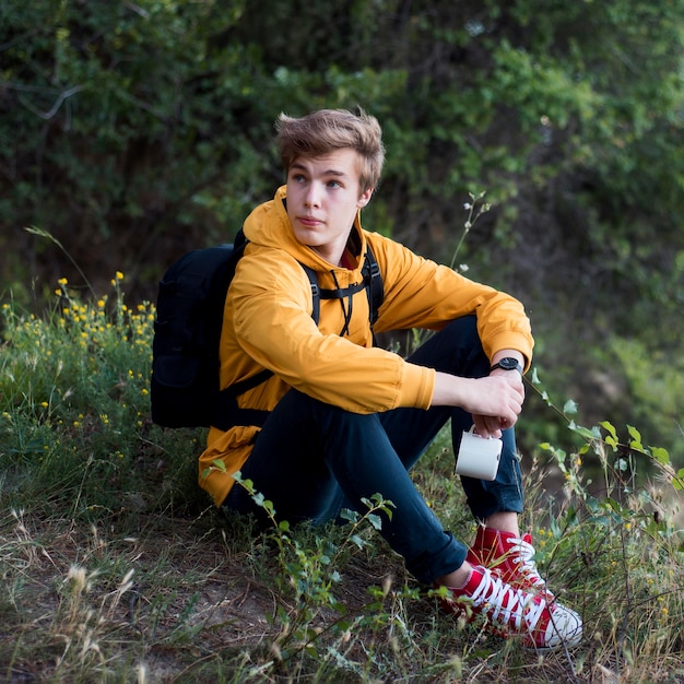 Volledige shot tiener met rugzak zittend op de grond in het bos