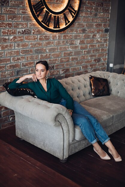 Volledige stockfoto van prachtige brunette vrouw in casual elegante kleding en hoge hakken ontspannen op een klassieke bank met kussens en wegkijken. De kamer is in loftstijl.