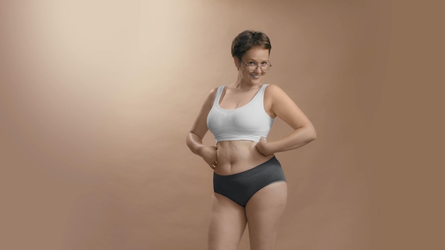Volslanke lichaamspositieve blanke vrouw in ondergoed kijkt in de camera glimlacht en is trots op haar vormen Beige studio achtergrond foto van hoge kwaliteit