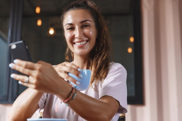 Volwassen blanke vrouw die 's ochtends thuis koffie drinkt, zittend op het terras met een kopje thee en een mobiele telefoon die gelukkig glimlacht naar de camera die tijd binnen doorbrengt