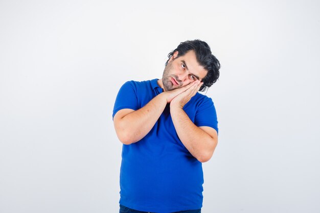 Volwassen man die op handpalmen als hoofdkussen in blauw t-shirt leunt en nadenkend kijkt. vooraanzicht.