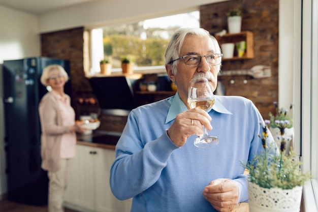 Gratis foto volwassen man die wijn drinkt en aan iets denkt terwijl zijn vrouw op de achtergrond eten aan het bereiden is