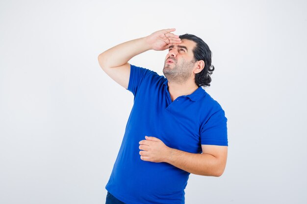 Volwassen man op zoek ver weg met hand boven het hoofd in blauw t-shirt, spijkerbroek en gefocust op zoek. vooraanzicht.