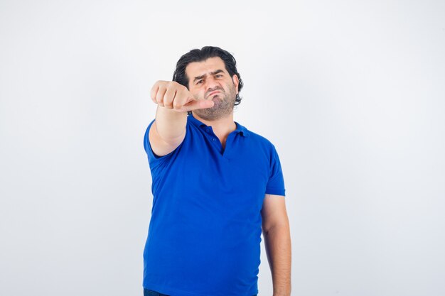 Volwassen man wijst opzij met duim in blauw t-shirt, spijkerbroek en kijkt teleurgesteld, vooraanzicht.