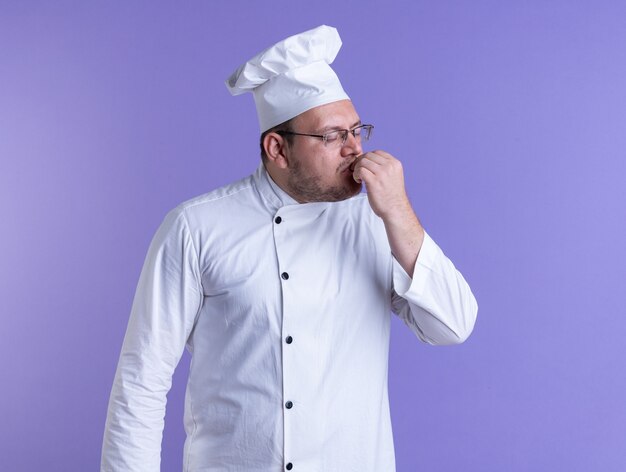 volwassen mannelijke kok met een uniform van de chef-kok en een bril die de lippen met de hand aanraakt met gesloten ogen geïsoleerd op een paarse muur