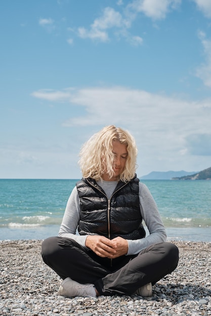 Volwassen toeristische europese blonde vrouw alleen zittend op een kiezelstrand met blauwe oceaan en lucht op de achtergrond verticaal schot Mensen en zomervakantie vakantie lifestyle concept