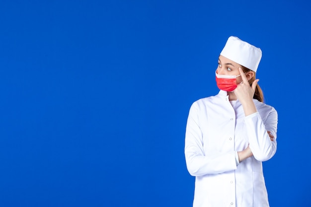 Vooraanzicht beklemtoonde jonge verpleegster in medisch kostuum met rood beschermend masker op blauwe muur