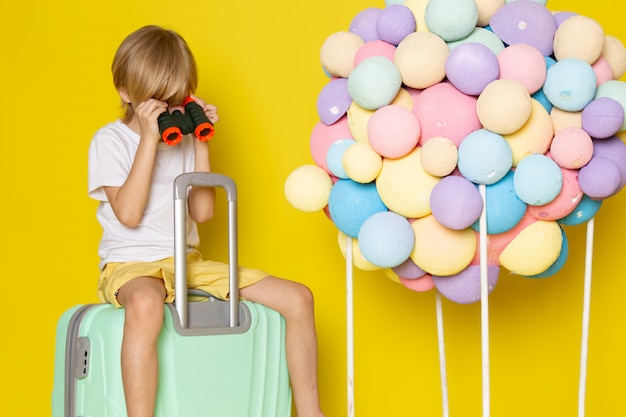 vooraanzicht blonde jongen in wit t-shirt zittend op de blauwe tas samen met veelkleurige lucht ballonnen op de gele vloer