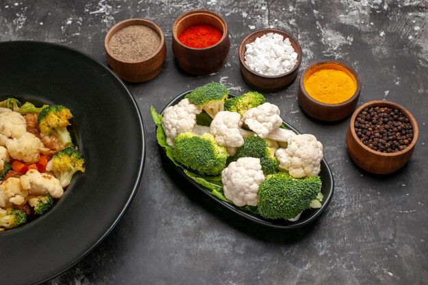 Vooraanzicht broccoli en bloemkoolsalade op zwarte ovale plaat verschillende kruiden in kleine kommen rauwe broccoli en bloemkool op donkere achtergrond