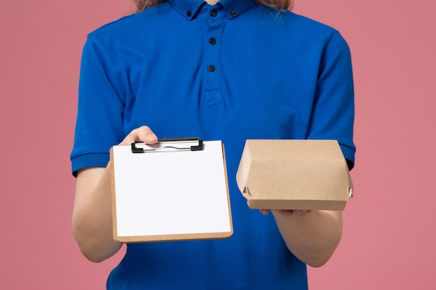 Vooraanzicht close-up vrouwelijke koerier in blauw uniform en cape met een klein voedselpakket voor bezorging met blocnote op roze muur, baanbezorgingsmedewerker werk