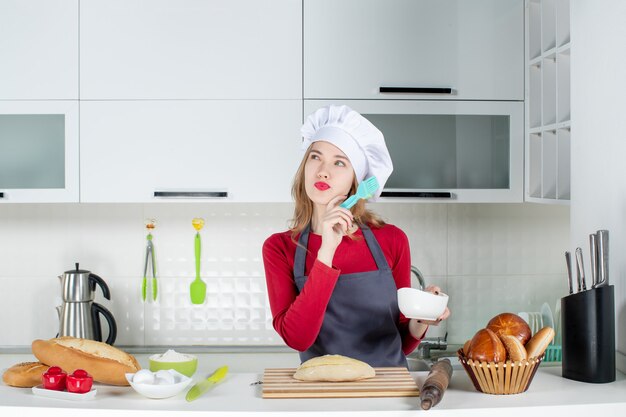 Vooraanzicht denkende blonde vrouw in koksmuts en schort die brood in de keuken botert