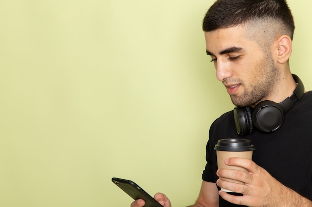 Vooraanzicht dichtbij jong aantrekkelijk mannetje in de zwarte telefoon van de t-shirtholding en het luisteren aan de koffiekop van de muziekholding