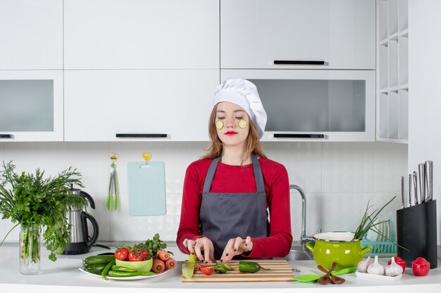 Vooraanzicht drukke vrouwelijke chef-kok in koksmuts die plakjes komkommer op haar gezicht zet