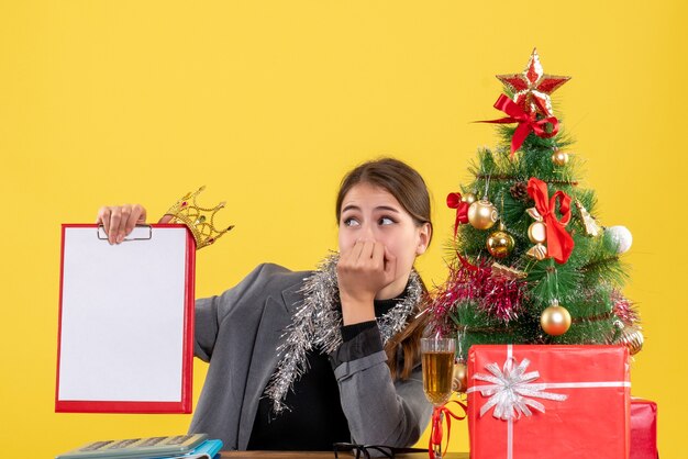 Vooraanzicht dwaalde meisje zittend aan de tafel met document met kroon kerstboom en geschenken cocktail