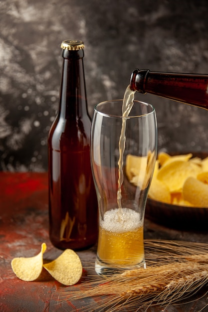 Vooraanzicht fles beer die in het glas stroomt met cips op de donkere achtergrond