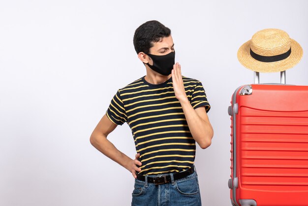 Vooraanzicht geeuwende jonge toerist met zwart masker dat zich dichtbij rode koffer bevindt
