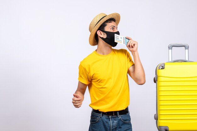 Vooraanzicht geïnteresseerde mannelijke toerist in gele t-shirt die zich dichtbij gele koffer bevindt die reiskaartje steunt