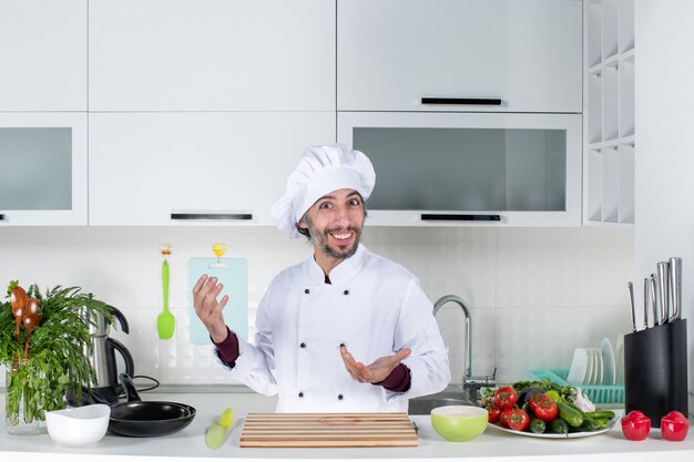 Vooraanzicht gelukkige mannelijke chef-kok in koksmuts die achter de keukentafel in de keuken staat
