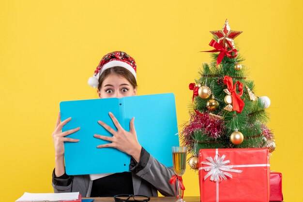 Vooraanzicht geschokt meisje met kerstmuts zittend aan de tafel die haar gezicht bedekt met de kerstboom van de documentmap en geschenkencocktail