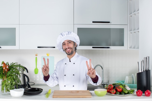 Vooraanzicht glimlachende mannelijke chef-kok in kokshoed die een overwinningsteken maakt dat achter de keukentafel staat