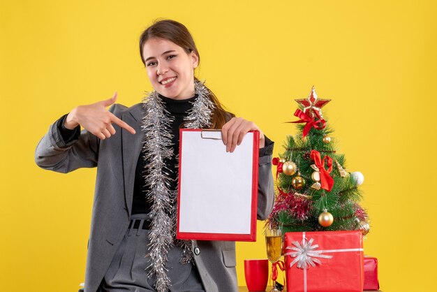 Vooraanzicht glimlachte meisje dat documenten toont die zich dichtbij Kerstmisboom en giftencocktail bevinden