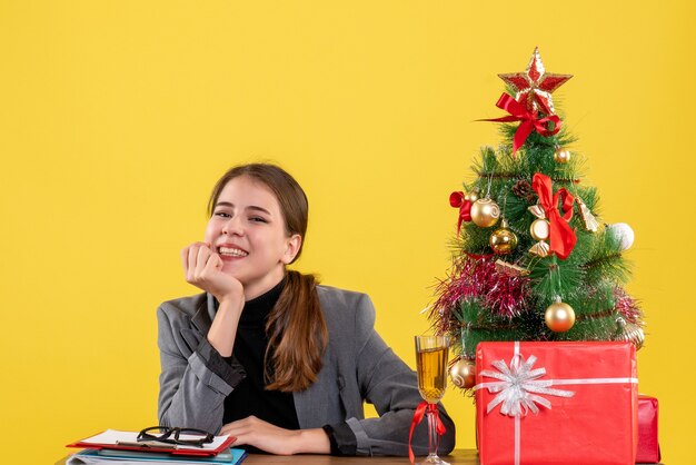 Vooraanzicht glimlachte meisjeszitting bij het bureau die hand aan haar kaakkerstmisboom en giftencocktail brengen