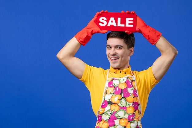 Gratis foto vooraanzicht grappige mannelijke huishoudster in geel t-shirt met verkoopbord boven zijn hoofd op blauwe ruimte