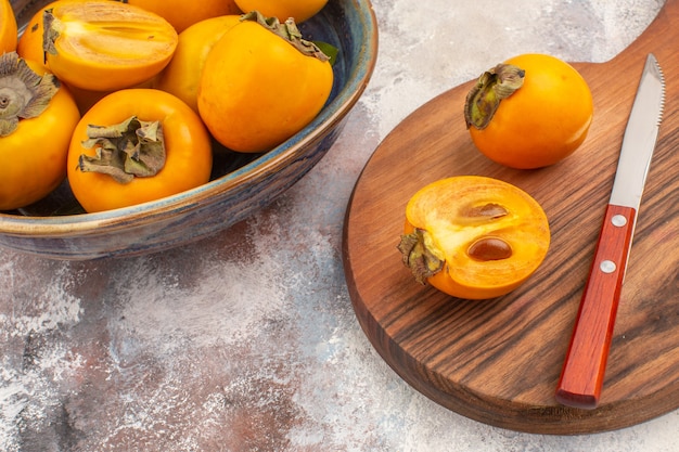 Gratis foto vooraanzicht heerlijke dadelpruimen in een kom persimmon en mes op snijplank op naakte achtergrond