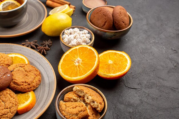 Vooraanzicht heerlijke zandkoekjes met verse sinaasappels en kopje thee op een donkere achtergrond, fruitkoekje, zoete koekje citrus